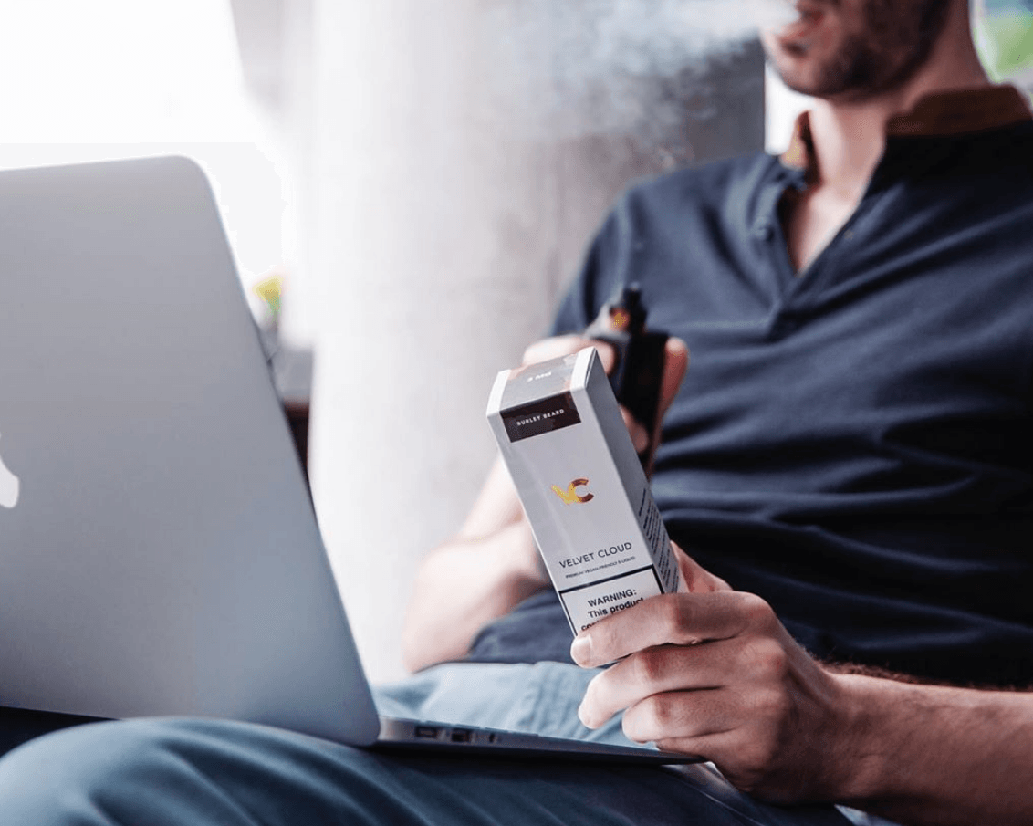 young man holding Velvet Cloud e-liquid vape juice with a vape while looking at his laptop