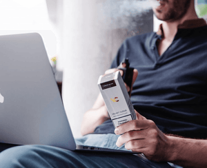 young man holding Velvet Cloud e-liquid vape juice with a vape while looking at his laptop 